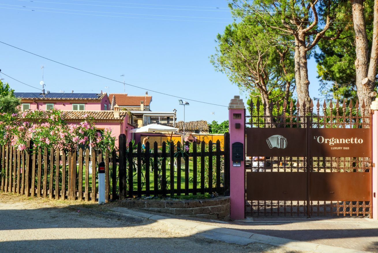 A gated entrance with a sign reading 'Organetto Luxury B&B' and a wooden fence with pink flowering plants.