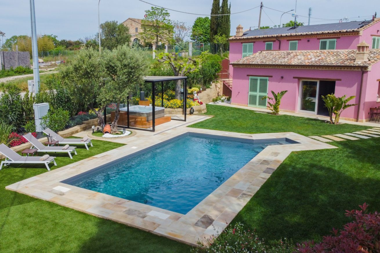 Une piscine bordée de pierres, entourée d’une pelouse bien entretenue et d’un bâtiment rose aux volets verts.