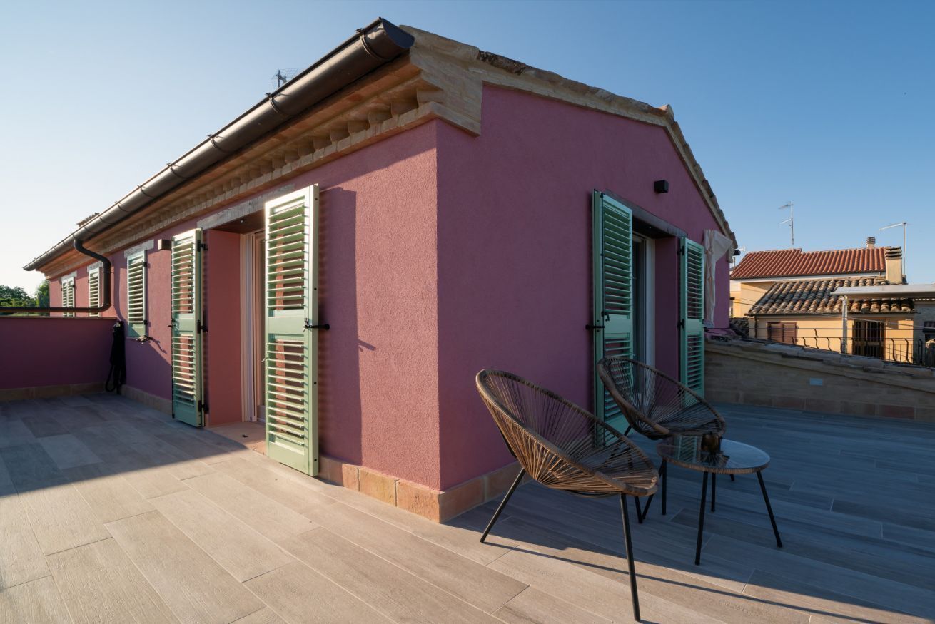 A pink building with green shutters, a tiled roof, and a patio with chairs and a table.