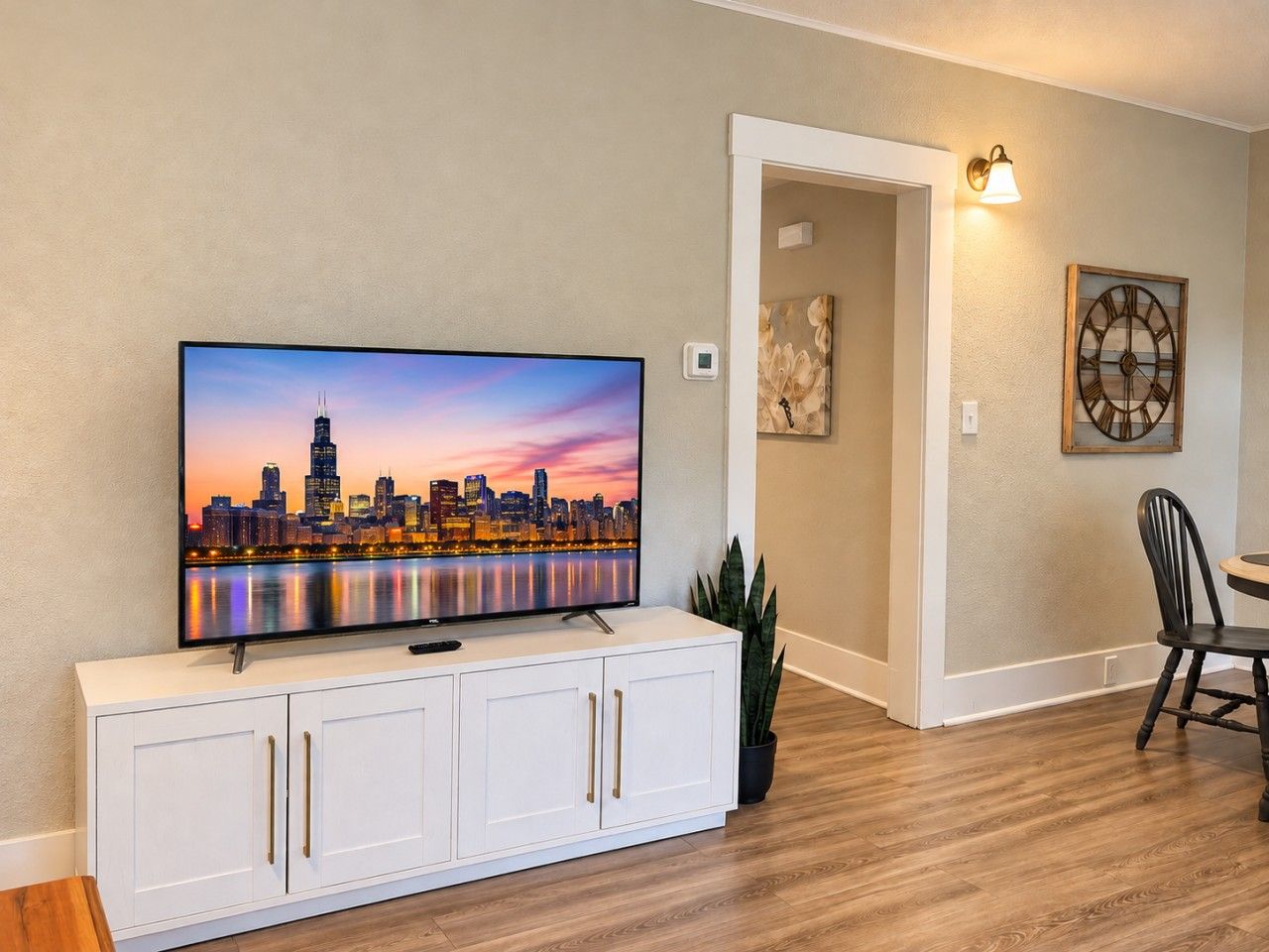 Un televisor de pantalla plana montado en un mueble blanco, un comedor con mesa y sillas, y un reloj de pared.