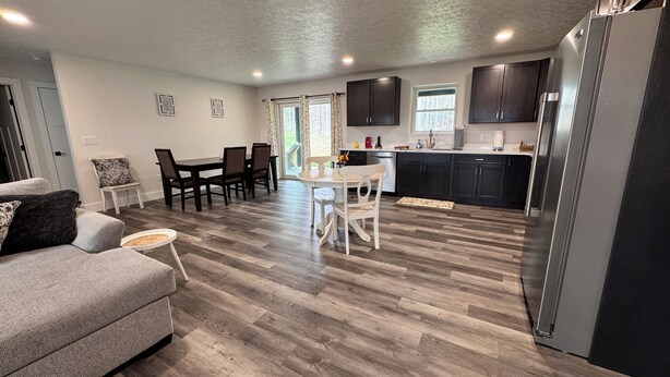 A modern kitchen with dark cabinets, a stainless steel refrigerator, and a dining area with a table and chairs. - Close to everything-tucked away from the noise. A brand-new, blissful stay. (Ithaca)