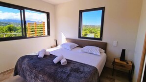A bedroom with a bed, two windows, a nightstand, and a view of greenery. - Villa Florida, Pinarello, 8 pers, piscine chauffée, 350 m plage à pieds (STE LUCIE DE PORTO VECCHIO)