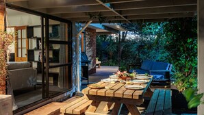 An outdoor dining area with a wooden table and benches, surrounded by greenery and a brick wall. - Comfort, Style, Relaxation. Your Home Away from Home Awaits! (Blenheim)