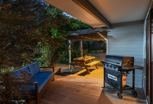 A covered outdoor patio with a wooden deck, a blue sofa, a picnic table, a grill, and a cart labeled "jumbuuk." - Comfort, Style, Relaxation. Your Home Away from Home Awaits! (Blenheim)