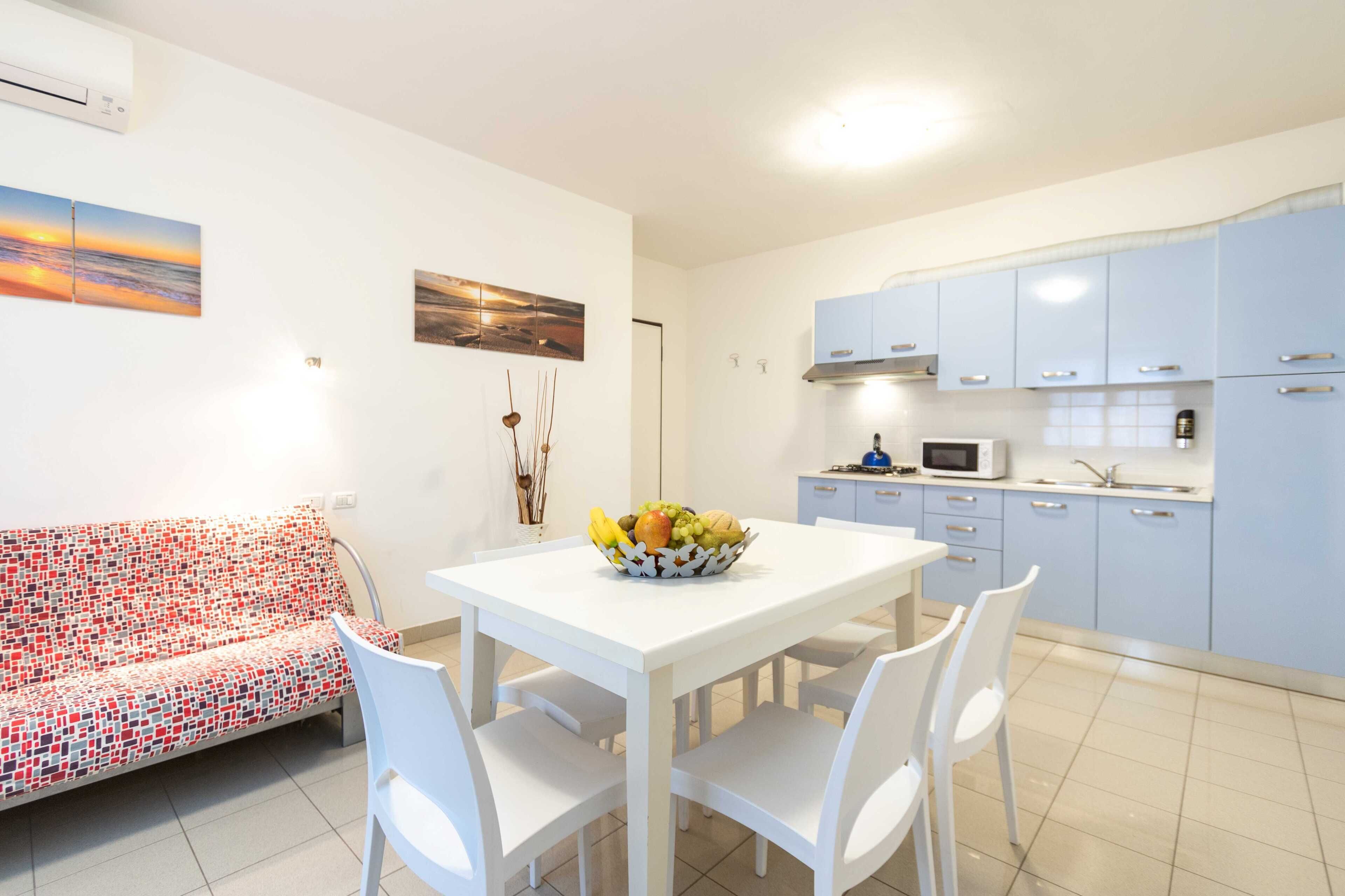 Una cocina compacta con armarios blancos, un microondas y un lavamanos. Una mesa de comedor con sillas se encuentra frente a la cocina.