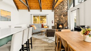 A cozy living area with a stone fireplace, wooden ceiling beams, and a dining table with chairs. - EV Charger, Dogs welcome at Sunset Ridge! (Lake Arrowhead)