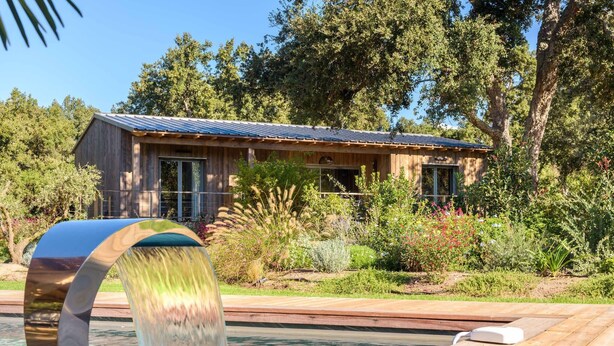 Una cabaña de madera con techo metálico, rodeada de exuberante vegetación y una pequeña piscina con una fuente de agua moderna.