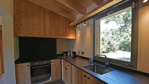 Cocina moderna con armarios de madera, horno empotrado, ventana grande con vistas a árboles y pileta de acero inoxidable.