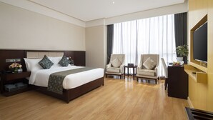 Habitación de hotel con una cama grande, dos sillones, televisión y un ventanal con cortinas.