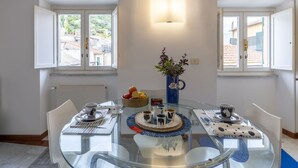 A dining area with a glass table, white chairs, a vase with flowers, and a basket of fruit. - Lovely apartment for 5 people with TV (Noli)