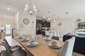 A modern dining area with a long table set for six, a kitchen in the background, and a living area with a sofa and TV.
