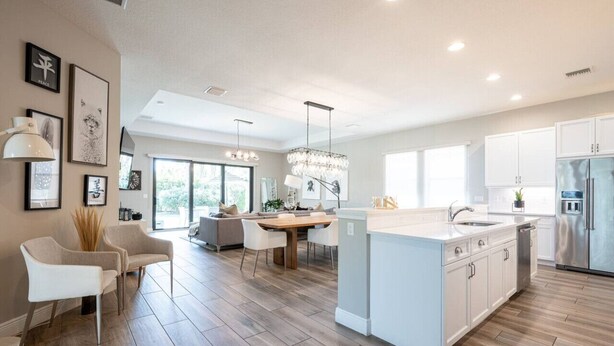 A modern kitchen with white cabinetry, a central island, and a dining area with a wooden table and chairs. - Luxurious Family Home - Spacious Kitchen & BBQ (Pompano Beach)