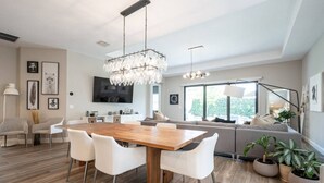 A modern living room with a wooden dining table, white chairs, a grey sofa, and a large window. - Luxurious Family Home - Spacious Kitchen & BBQ (Pompano Beach)