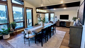 A modern dining area with a long wooden table, black chairs, and a white rug. There are large windows offering a view of a snowy landscape. - Winterfell at Winter Park Resort, New Luxury Home | Private Hot Tub | Stunning Views (Winter Park)
