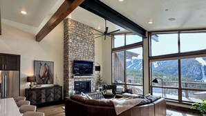 A spacious living room with a stone fireplace, a large TV mounted on the wall, and a view of mountains through large windows. - Winterfell at Winter Park Resort, New Luxury Home | Private Hot Tub | Stunning Views (Winter Park)