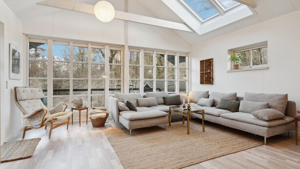 A spacious living room with a large sectional sofa, a chaise lounge, and a coffee table. There are large windows providing ample natural light and a view of the outdoors.