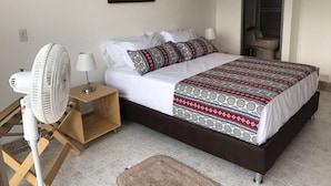 Ein ordentlich bezogenes Bett mit gemusterten Betttextilien, ein Standventilator, eine Nachttischlampe und ein Nachttisch aus Holz.