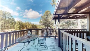 A covered outdoor patio with a table and chairs, surrounded by a wooden railing, overlooking a forested area. - Close to Lifts | WIFI | Easy Access | Pinetree 318 (Angel Fire)