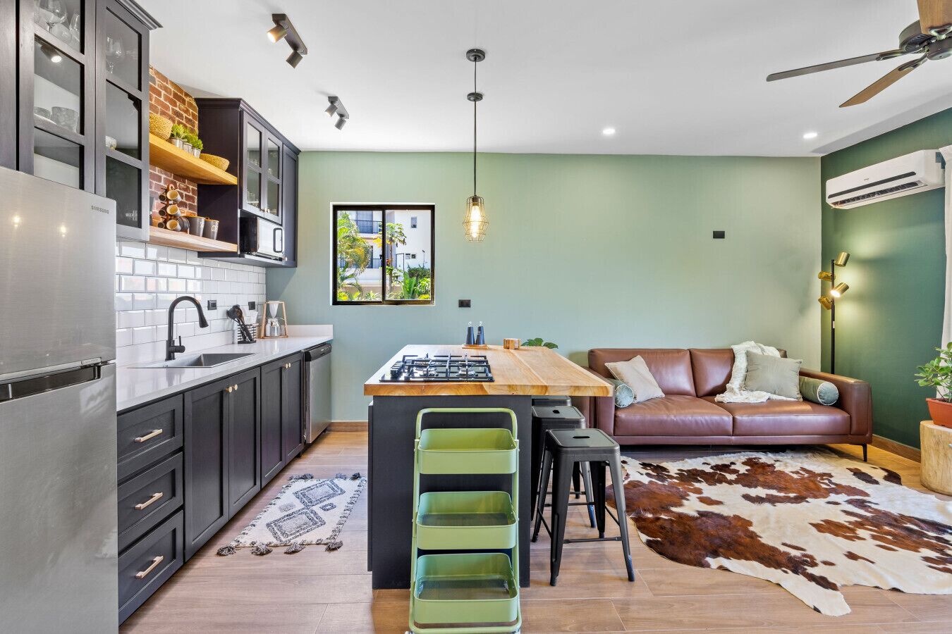 Une cuisine moderne avec un îlot central, des meubles de couleur sombre et un canapé vert dans un espace de vie agrémenté d’un tapis en peau de vache.
