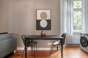 Un coin repas avec une table, des chaises et un vase. Il y a une fenêtre avec des rideaux et un radiateur.