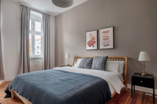 Une chambre à coucher comprenant un lit, deux tables de chevet, une fenêtre avec des rideaux et deux œuvres d’art accrochées au mur.