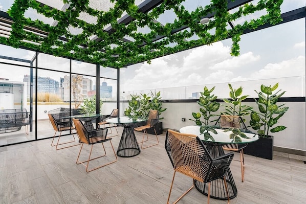 A modern rooftop terrace with glass walls, furnished with round tables and chairs, and adorned with potted plants.