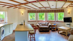 Un amplio espacio de sala con cocina, vigas de madera en el techo, ventanas grandes, un sofá y un televisor.
