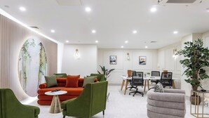 Ein moderner Arbeitsraum mit einem roten Sofa, grünen Sessel und einem grauen Sessel. Es gibt Schreibtische mit Computern und Stühlen sowie eine große Pflanze in der Ecke.