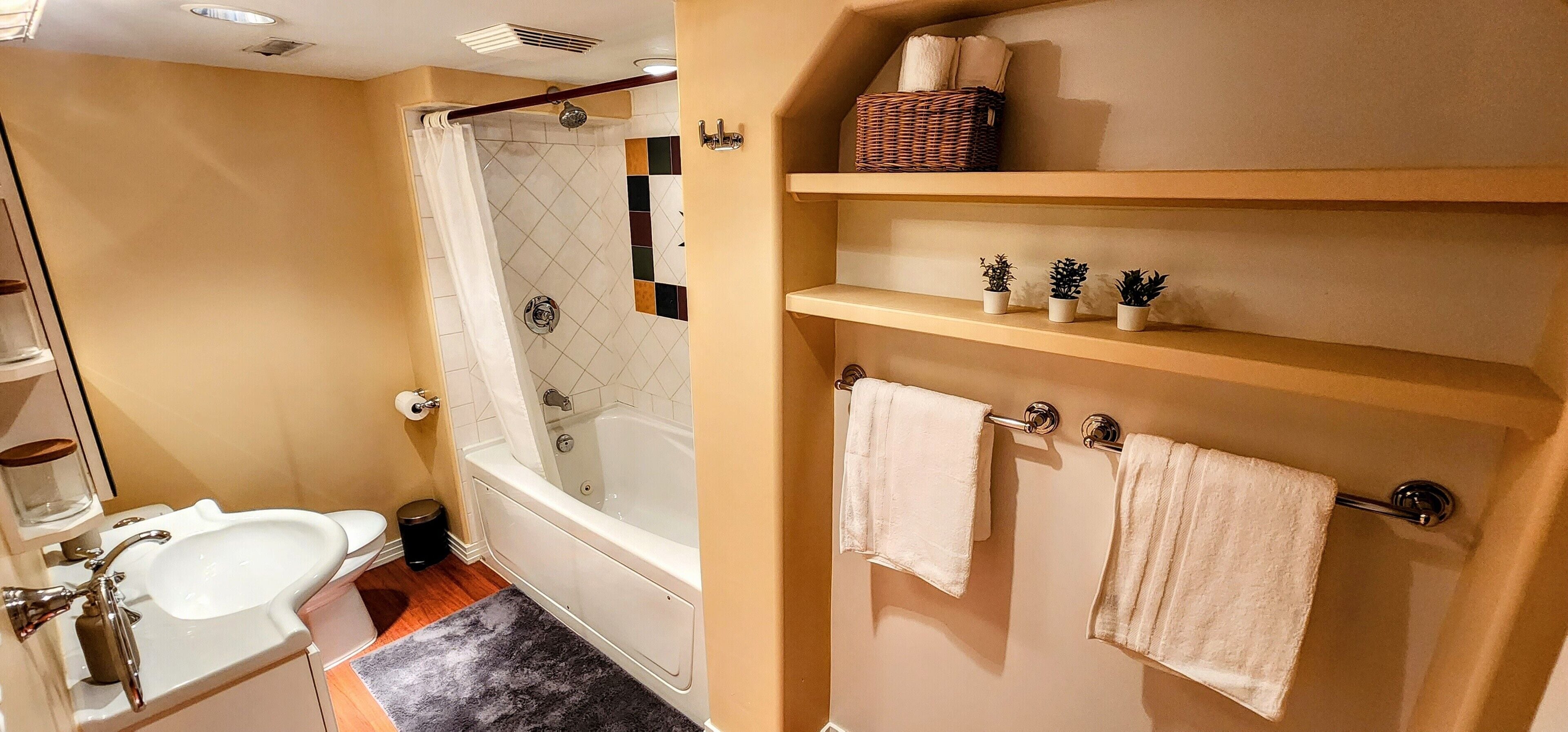 A bathroom with a bathtub, a sink, a shower, and shelves with towels and potted plants.