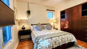 A bedroom with a bed, bedside lamps, a hanging plant, a window with blinds, and a wardrobe with drawers.