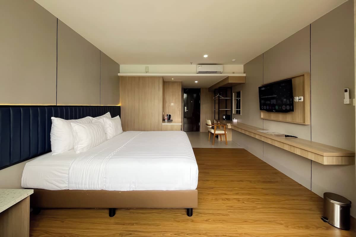 A modern hotel room with a large bed, a desk, a chair, a TV mounted on the wall, and a trash bin.