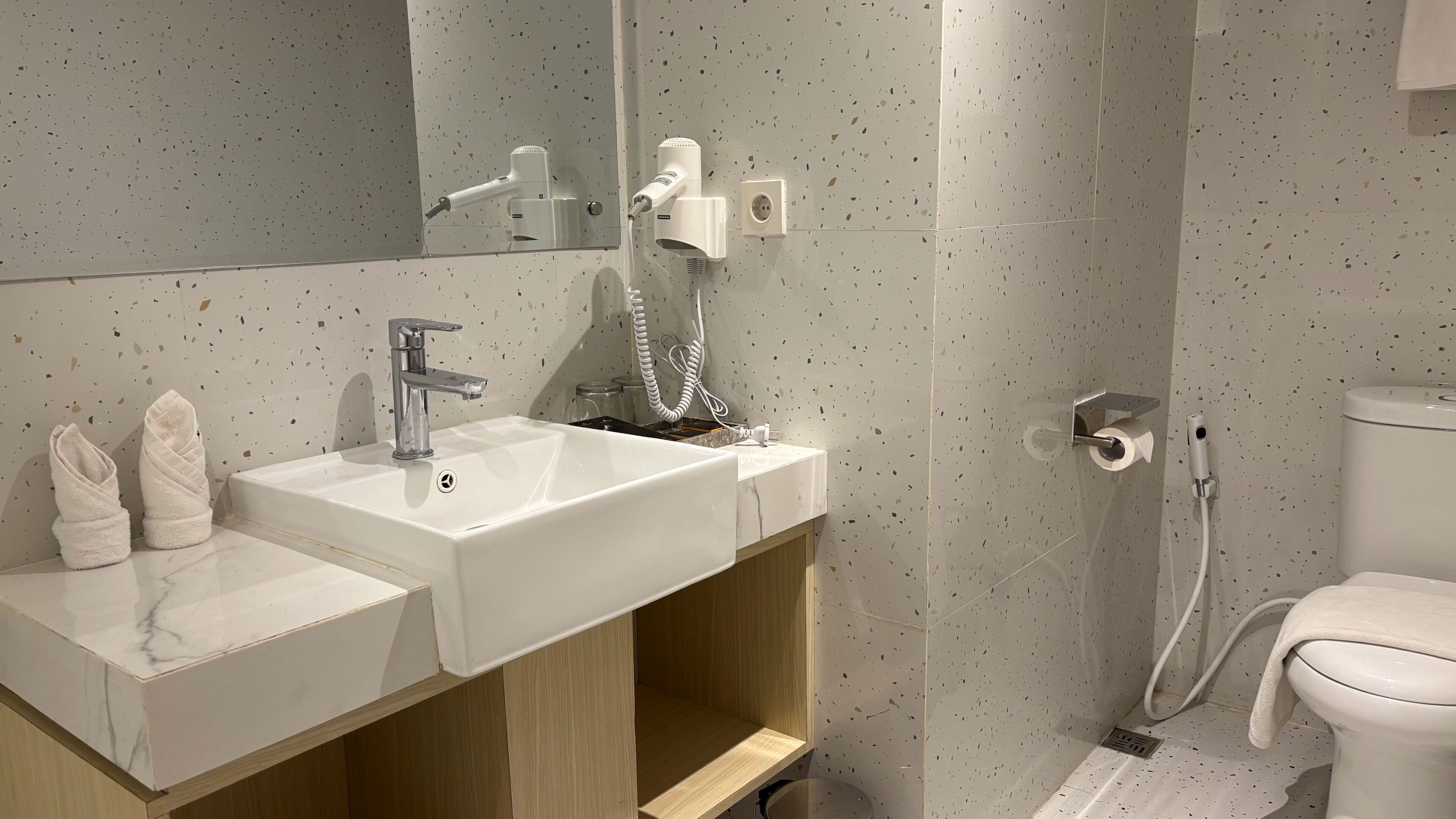 Un baño moderno con lavamanos, espejo y inodoro blancos. Hay dos toallas sobre el mostrador del lavamanos, y un secador de pelo está montado en la pared.