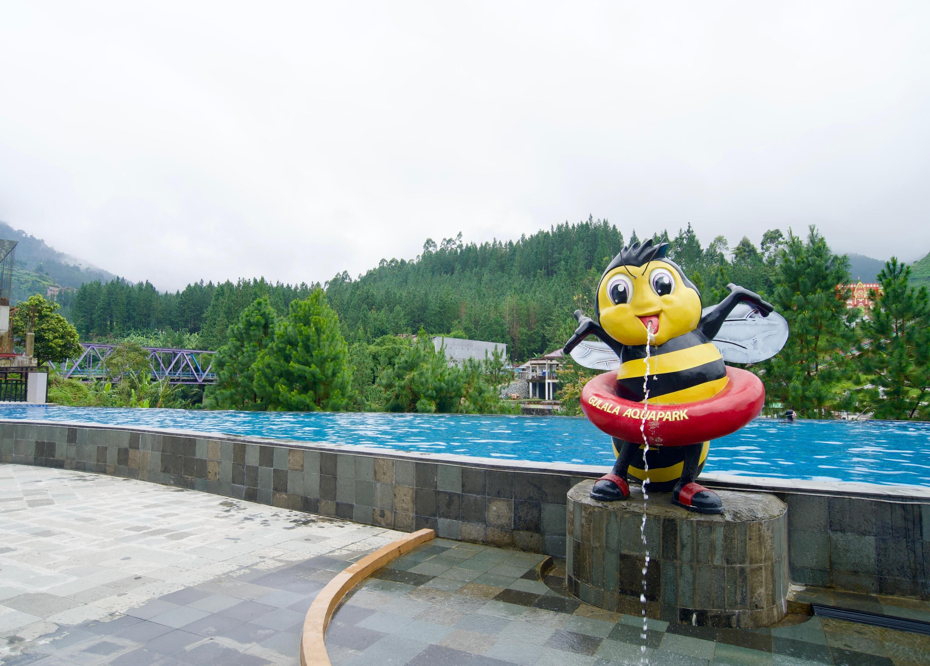 En stor bi-statue med en rød og gul flyer med teksten "Gulala Aquapark" ved en swimmingpool.