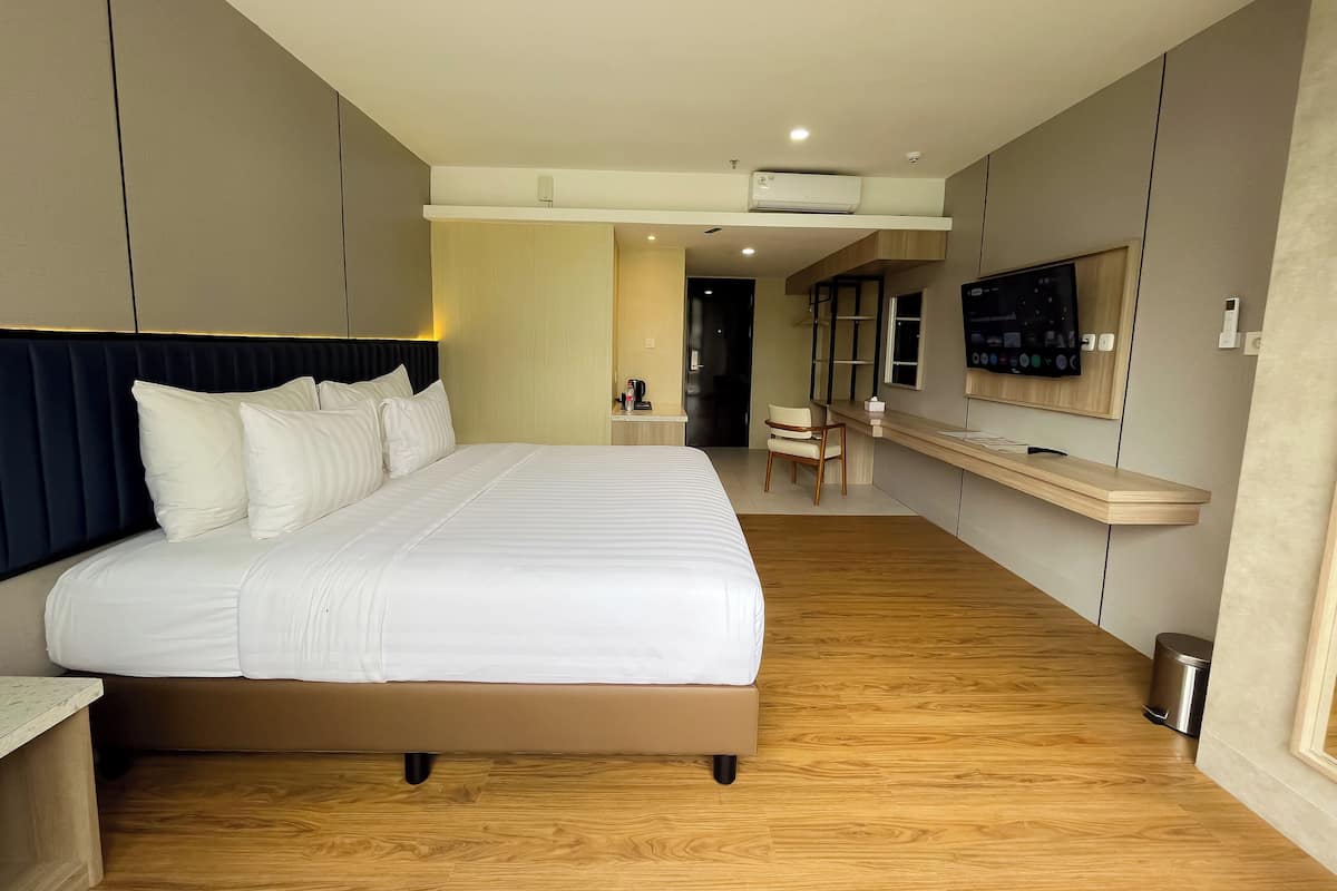 A modern hotel room with a large bed, a desk, a chair, a TV, and a trash bin.