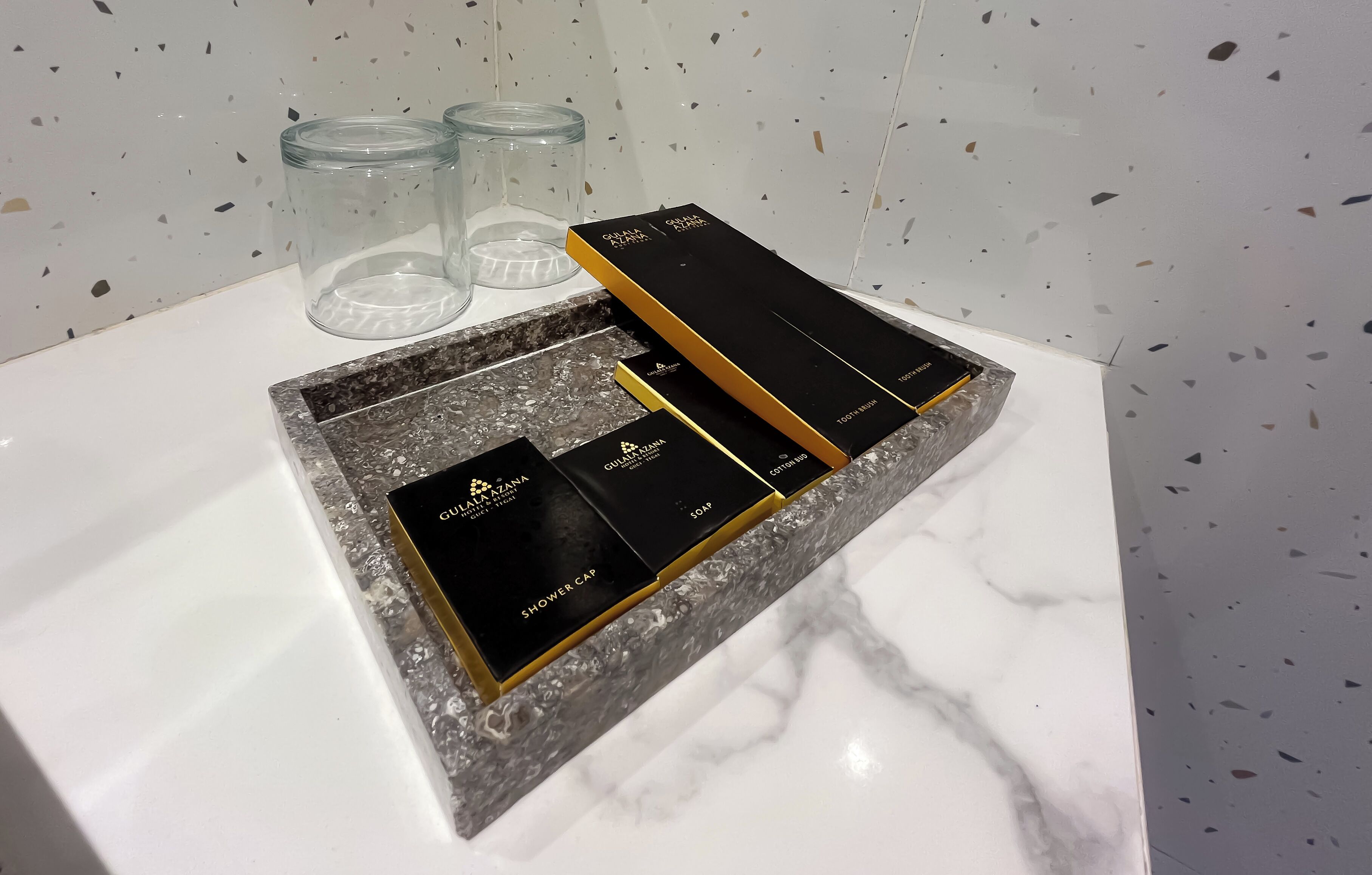 Un meuble-lavabo de salle de bain avec un plan de toilette en marbre, un bonnet de douche noir et doré, et une boîte noire portant l’inscription « Gulshan Azam ».