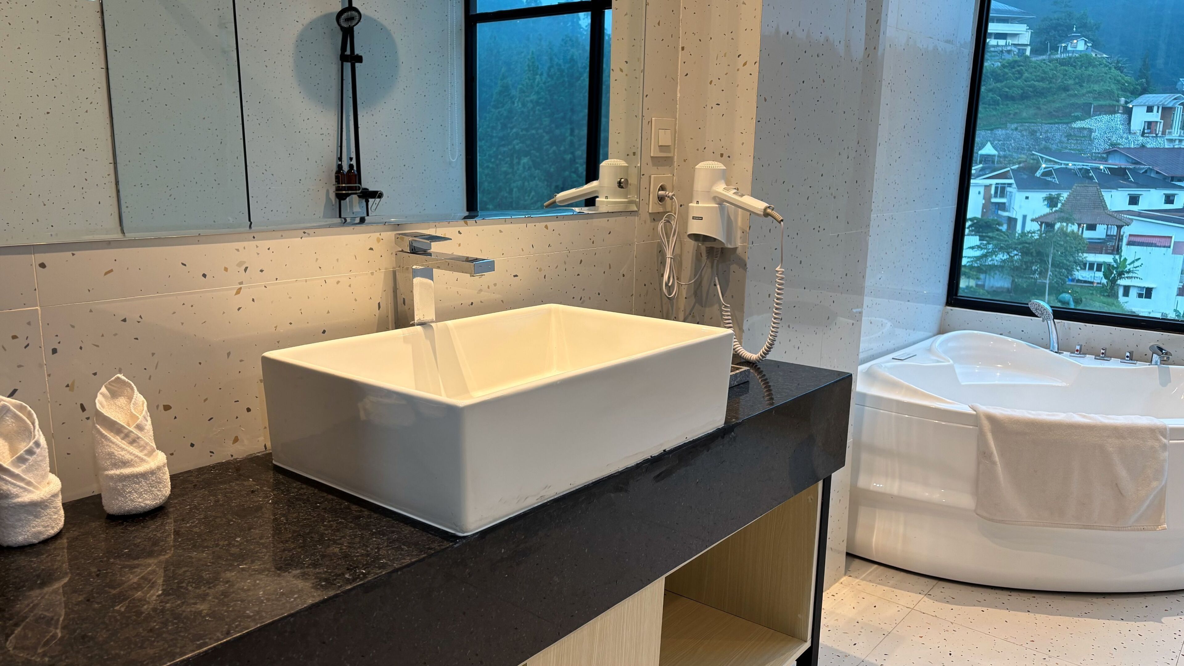 A modern bathroom with a large rectangular sink, a bathtub, and a window offering a view of a residential area.