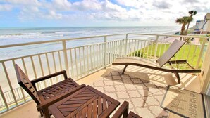 Een balkon met uitzicht op zee, een houten tafel, twee lounge fauteuils en een vloerkleed met dessin.