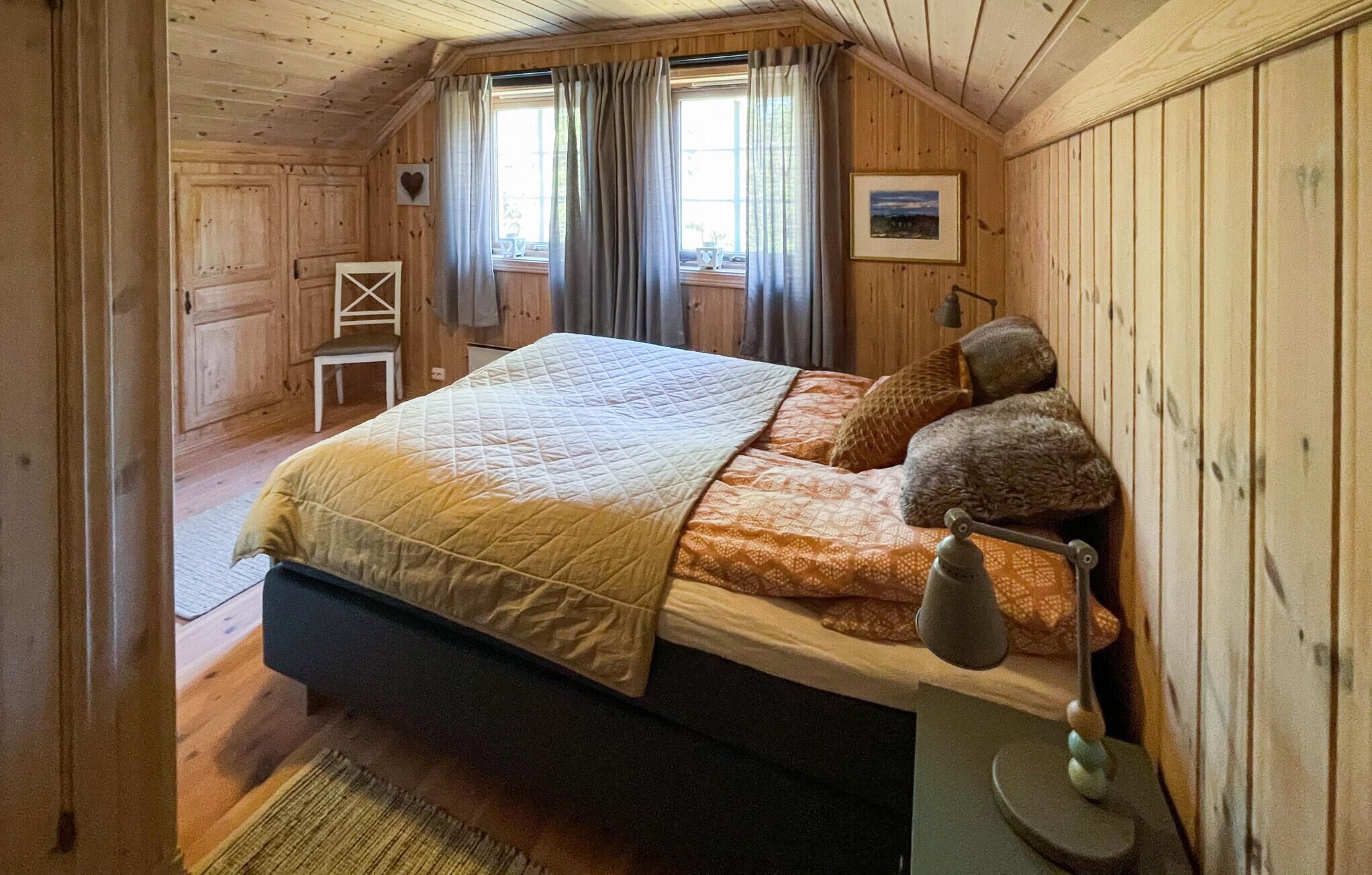 Een gezellige slaapkamer met een houten plafond, een bed met een gewatteerde sprei, twee kussens, een bedlamp en een kleine stoel.