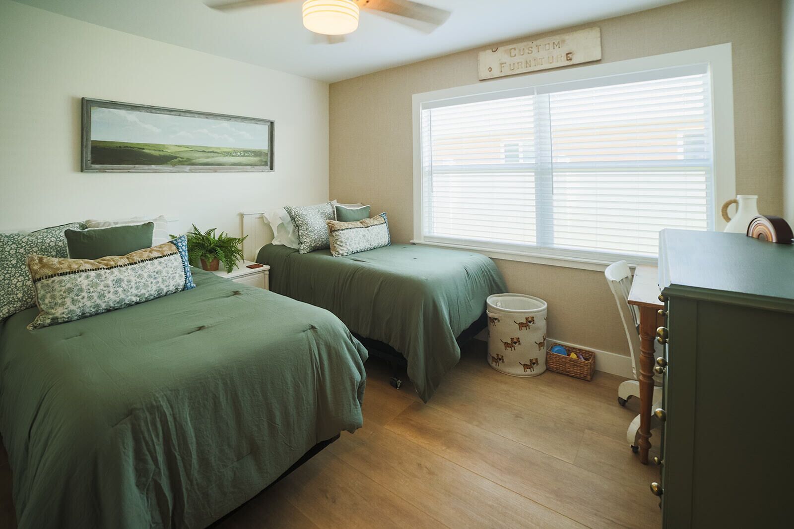 A bedroom with two beds, a window with blinds, a ceiling fan, a bedside table with a plant, and a dresser.