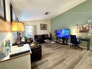 Een woonkamer met een leren bank, een televisie, een bureau met een stoel, en een prikbord aan de muur.