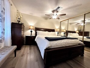 Een hotelkamer met een groot bed, twee nachtkastjes met lampen, een plafondventilator, een spiegel en een houten bankje.
