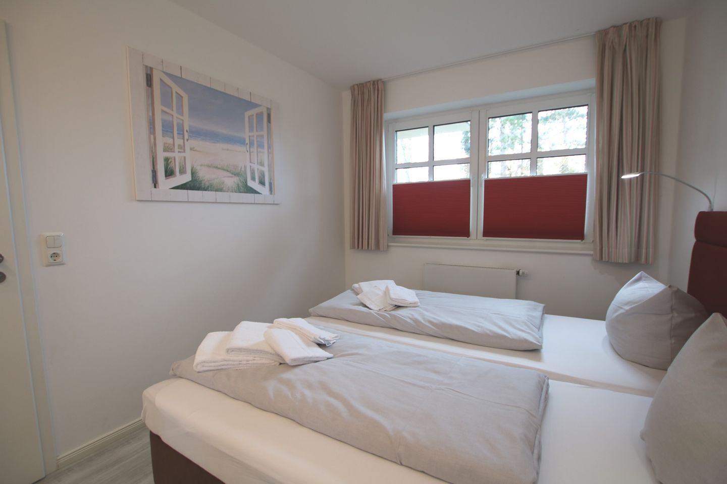 Habitación de hotel con dos camas, una ventana con persianas rojas, un cuadro con una playa y una lámpara.