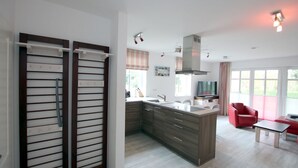 A modern kitchen with a central island, a dining area with a red chair, and a large window providing natural light. - Strandstraße 18c, Whg. 7, Haus Paul - Ferienwohnung mit 3- Zimmern auf ca. 70m² im EG mit Terrasse (Wyk auf Föhr)