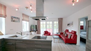 A modern kitchen with a central island, a dining area with a red sofa, and a separate living area with a red sofa and a TV. - Strandstraße 18c, Whg. 7, Haus Paul - Ferienwohnung mit 3- Zimmern auf ca. 70m² im EG mit Terrasse (Wyk auf Föhr)