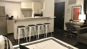 Ein kompaktes Hotelzimmer mit Küchenzeile, einem Essbereich mit Barhocker, einer kleinen Sitzecke mit einem Sessel und einem Bett mit gestreifter Bettdecke.