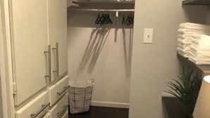 Ein begehbarer Kleiderschrank mit Bett im Hintergrund, einem Wäschekorb und einem Stapel gefalteter Handtücher.