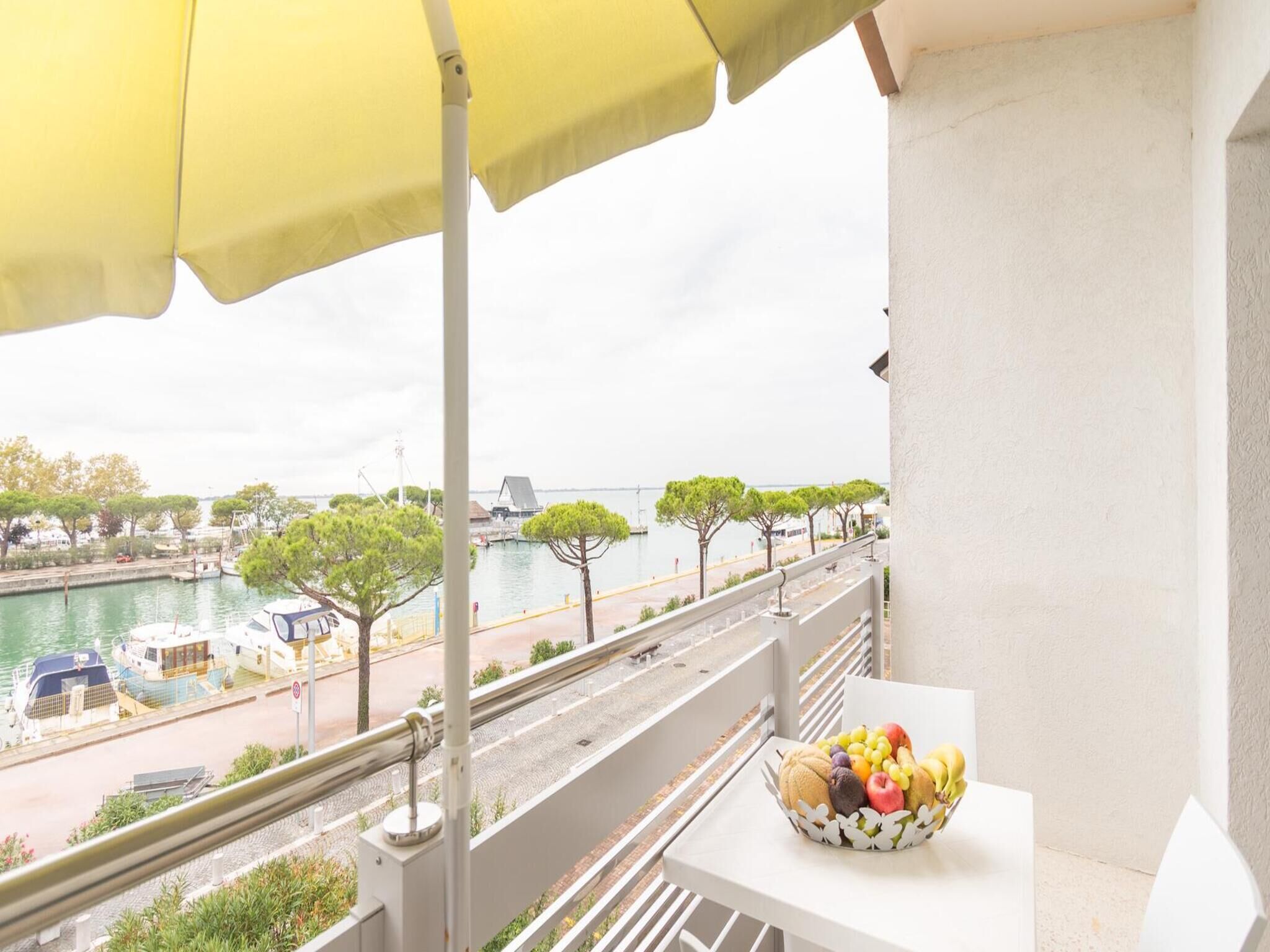 Ein Balkon mit einem gelben Sonnenschirm, einem Tisch mit Obstschale und Blick auf eine Marina mit Booten und Bäumen.