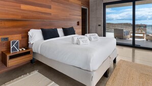 A modern bedroom with a large bed, wooden panel walls, a nightstand, and a view of the sea. - KARAT Villa Atelier de la Vega (Playa Paraiso)