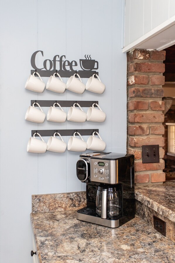 Una cocina con cafetera, un colgador de tazas en la pared y pared de ladrillo.