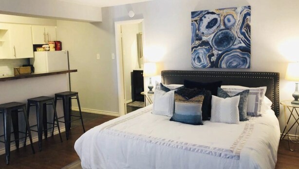 A bedroom with a large bed, a headboard, and a painting above it. There is a kitchen area with a refrigerator and a counter visible in the background. - Montrose Stylish Studio! Medical Center 45 (Houston)
